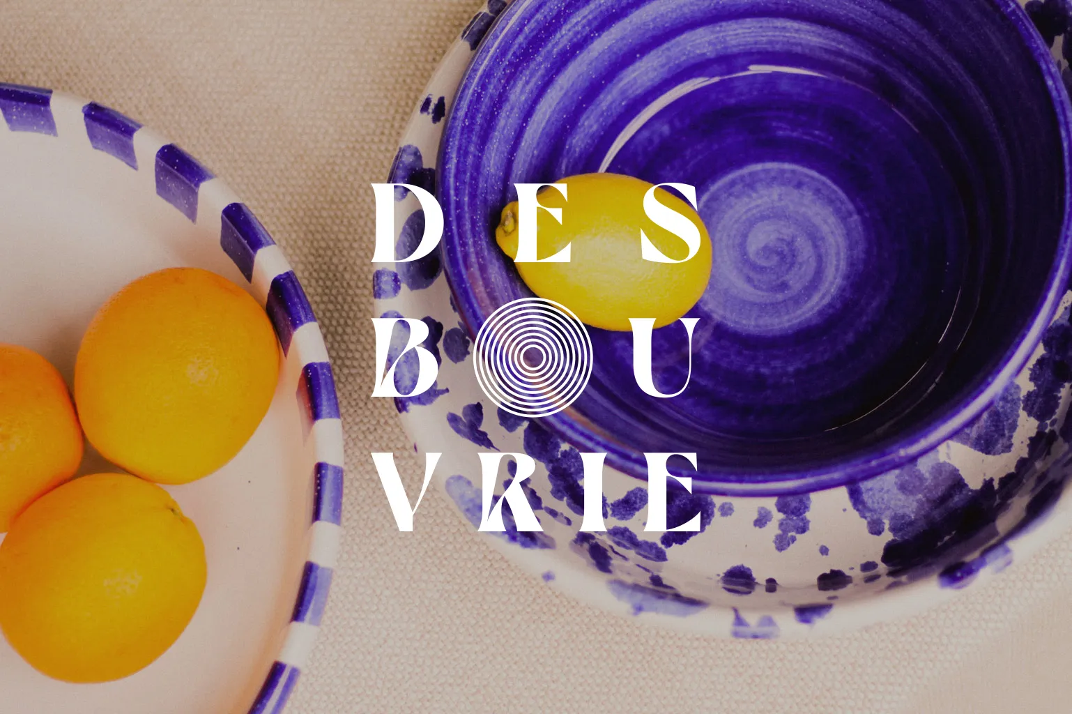 Des Bouvrie logo with its circular pictogram over blue glazed bowls holding oranges and a lemon