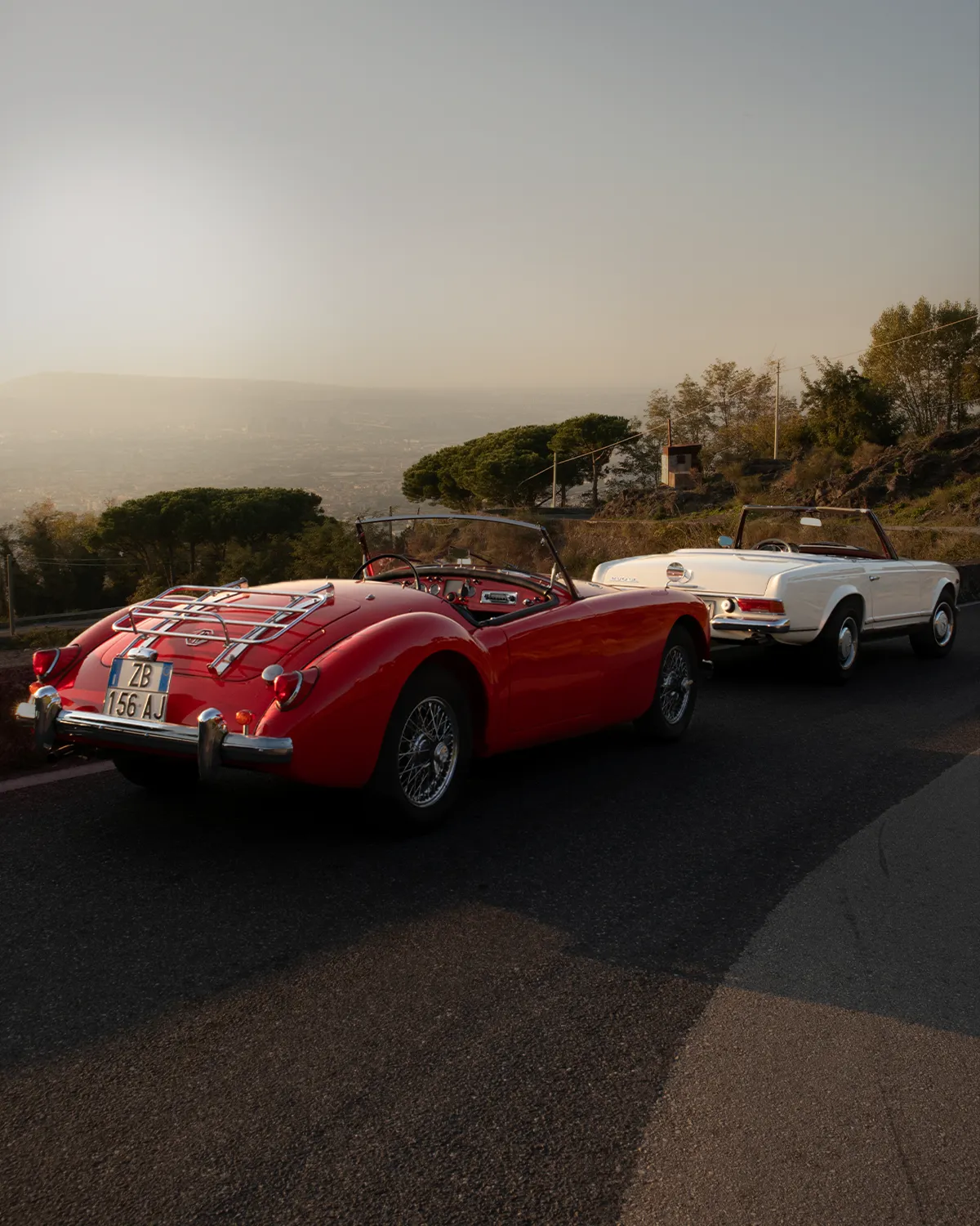 Red MGA and white Mercedes-Benz Pagoda convertibles on a hillside road at sunset