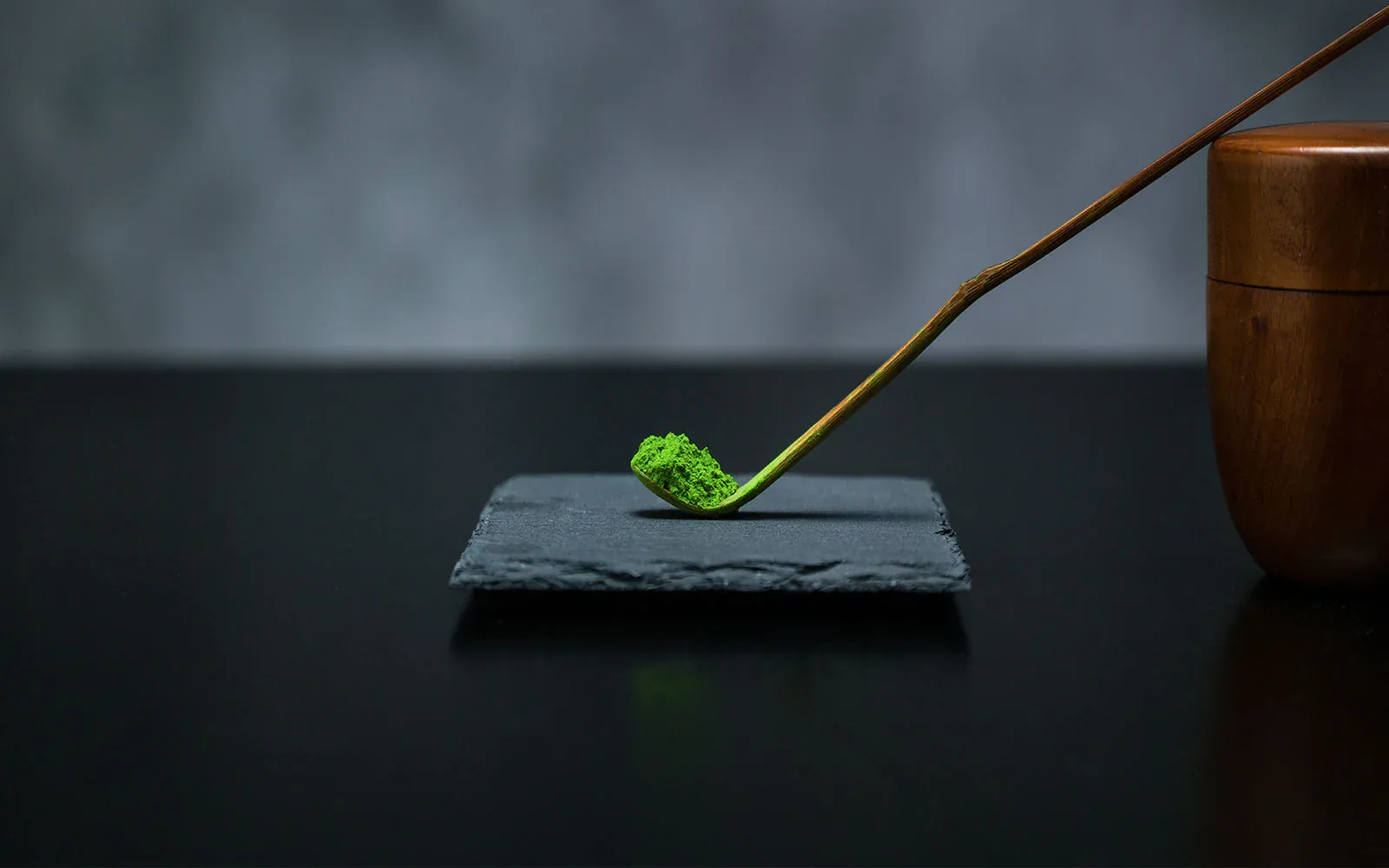 Bamboo scoop of matcha powder on a slate plate beside a wooden tea caddy